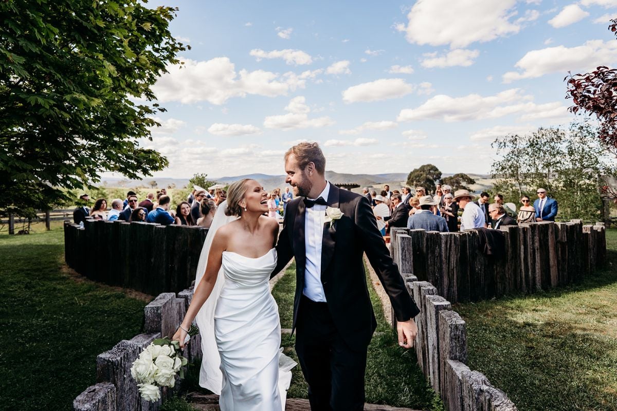 Eileen and Patrick smiling as they walk down the outdoor wedding aisle at Seclusions, Blue Mountains, with guests behind them.